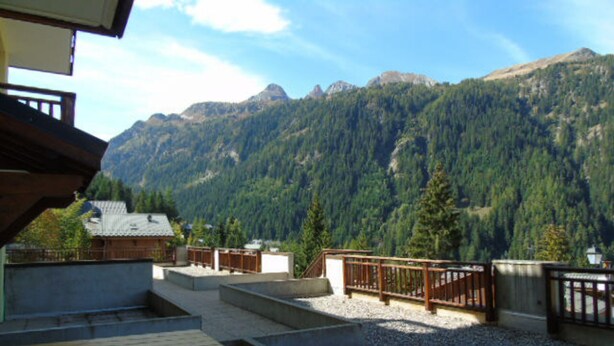 A terrace with a wooden table and benches, overlooking a mountainous landscape with dense forests and rocky peaks.
