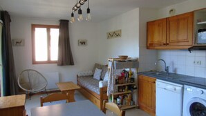 A compact kitchen with wooden cabinets, a washing machine, a dining table with chairs, a fan, and a small sofa.