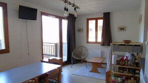 A room with a dining table, chairs, a fan, a television, and a shelf with various items.