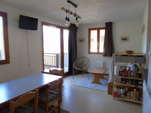Um quarto com uma mesa de jantar, cadeiras, um ventilador, uma televisão e uma estante com diversos objetos.