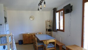 A compact kitchen with a dining table, chairs, a wooden cabinet, a clock, and a television mounted on the wall.