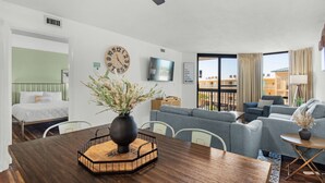 Ett modernt hotellrum med ett matbord i trä, en soffa, en TV och en balkong med utsikt.