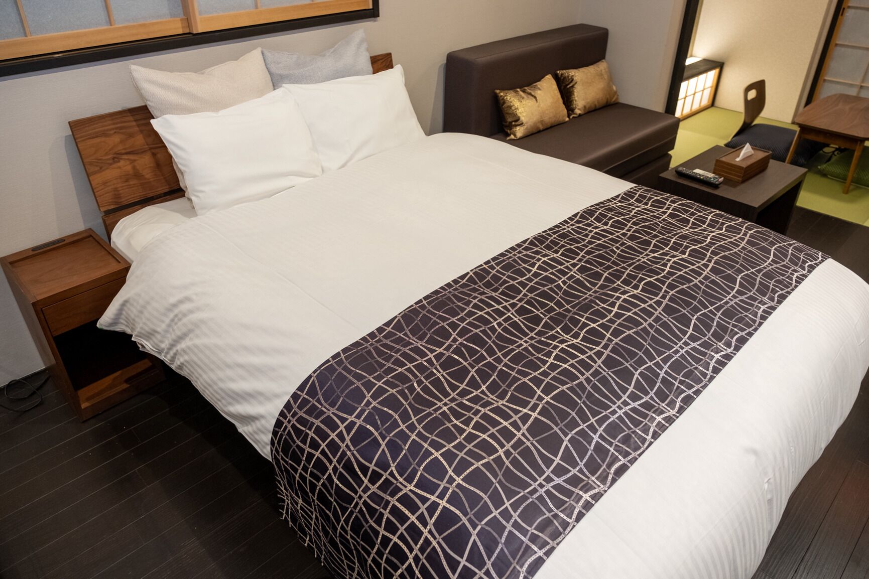 A neatly made bed with white and patterned bedding, a dark sofa with gold cushions, a small bedside table with a remote control, and a nightstand with a drawer.