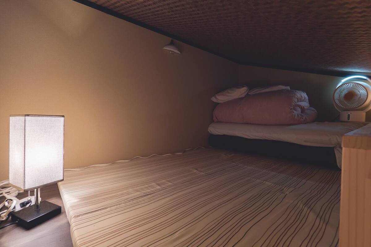 Une petite chambre agréable, dotée d’un lit simple, d’une table de chevet, d’une lampe et d’un ventilateur de plafond.