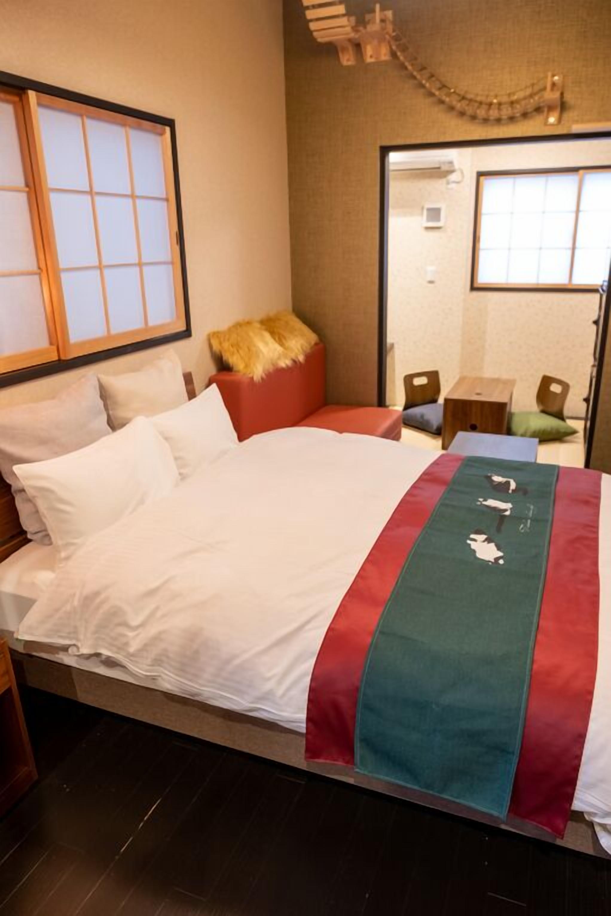 Quarto de hotel com cama, cabeceira de madeira, manta vermelha e verde, e uma pequena mesa de cabeceira de madeira.