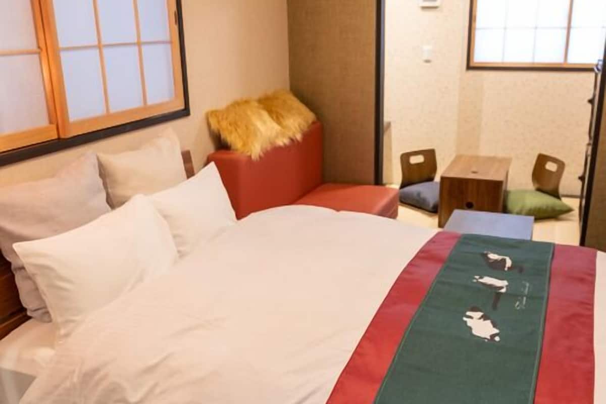 Une chambre d’hôtel avec un lit, une tête de lit en bois, un plaid rouge et vert, et une petite table de chevet en bois.