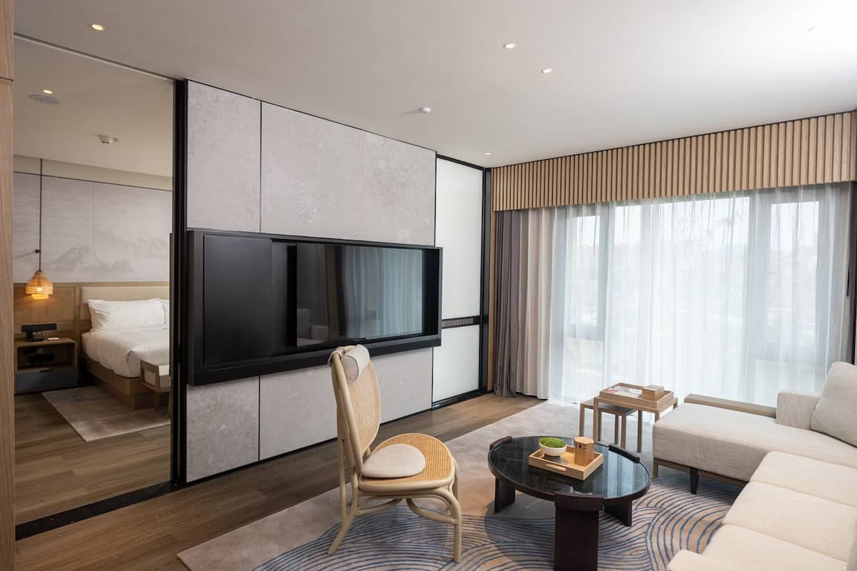 Une chambre d’hôtel moderne dotée d’un grand téléviseur à écran plat fixé au mur, d’un canapé confortable, d’une table à manger agrémentée d’un vase et d’une chaise en osier.