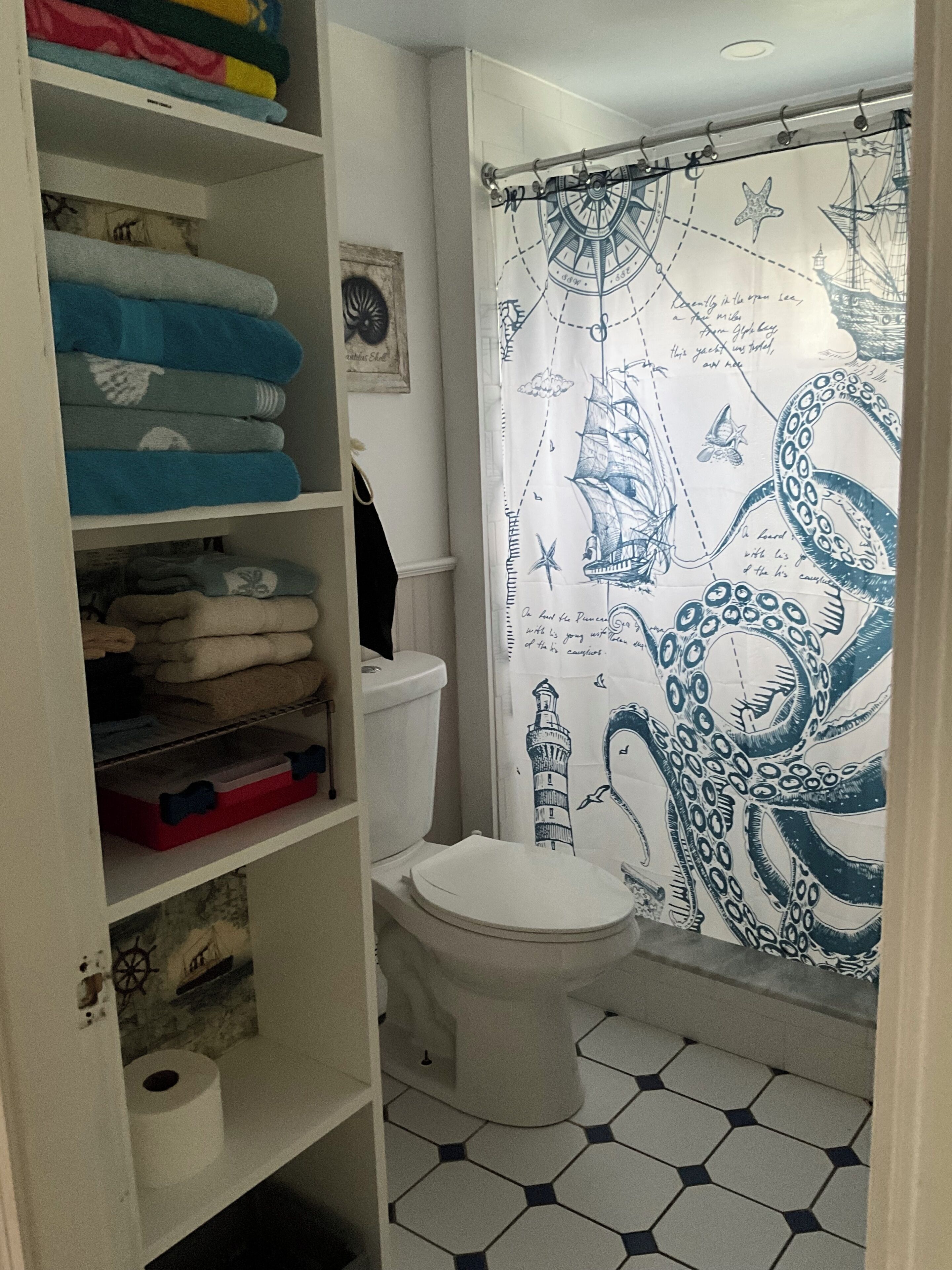 A bathroom with a white toilet, a shower curtain with nautical designs, and a shelf with folded towels.