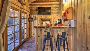 A wooden cabin interior with a stone fireplace, a bar area with stools, and a large window overlooking a stone exterior. - Greer Lodge- Guest Quarters (Greer)