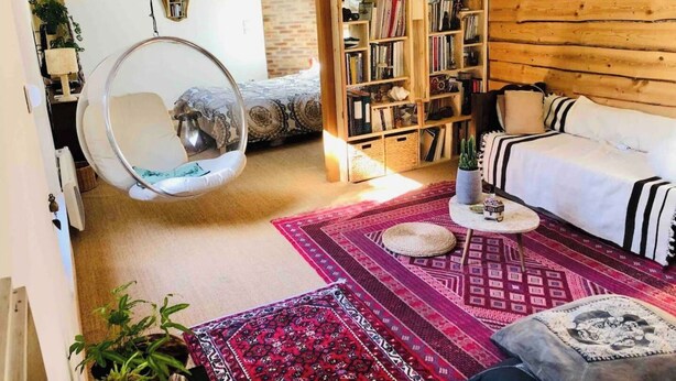 Een gezellige woonkamer met een hangstoel, een bank, een boekenkast en een kamerkleed met patroon.