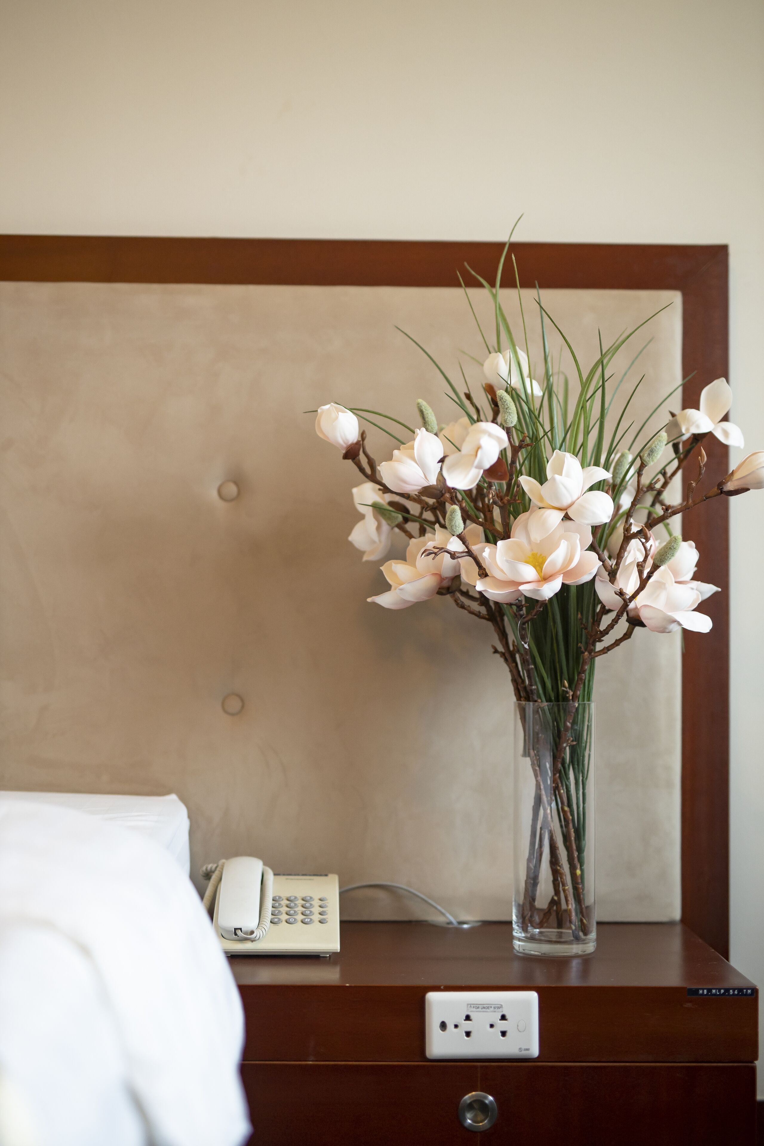 Ett hotellrum med en säng, ett sängbord, en telefon och en vas med blommor.