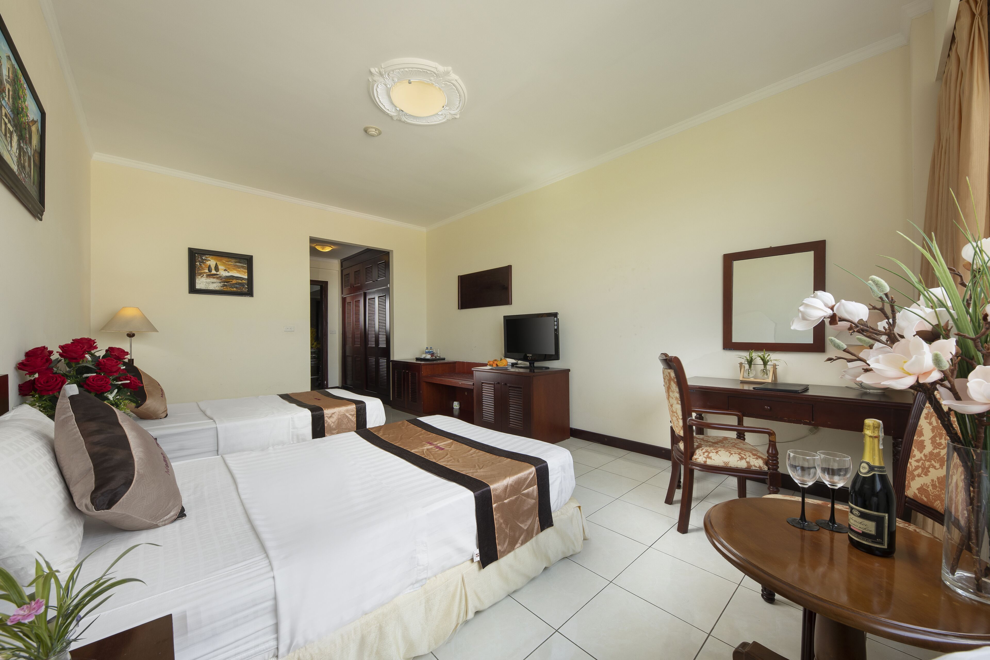 A hotel room with two beds, a desk with a chair, a television, and a dining table with two glasses and a bottle.