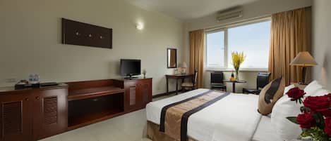A hotel room with a bed, a desk, a TV, a window with curtains, and a vase with flowers.