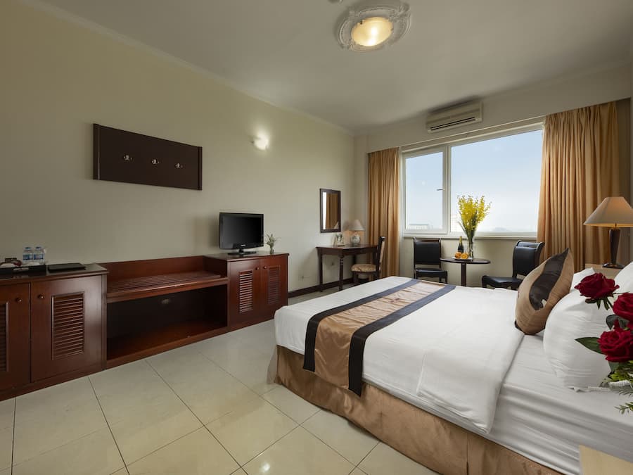 A hotel room with a bed, a desk, a TV, a window with curtains, and a vase with flowers.