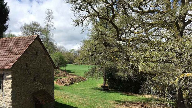 Un edificio de piedra con techo inclinado, un pequeño espacio de estar al aire libre y un campo verde al fondo.