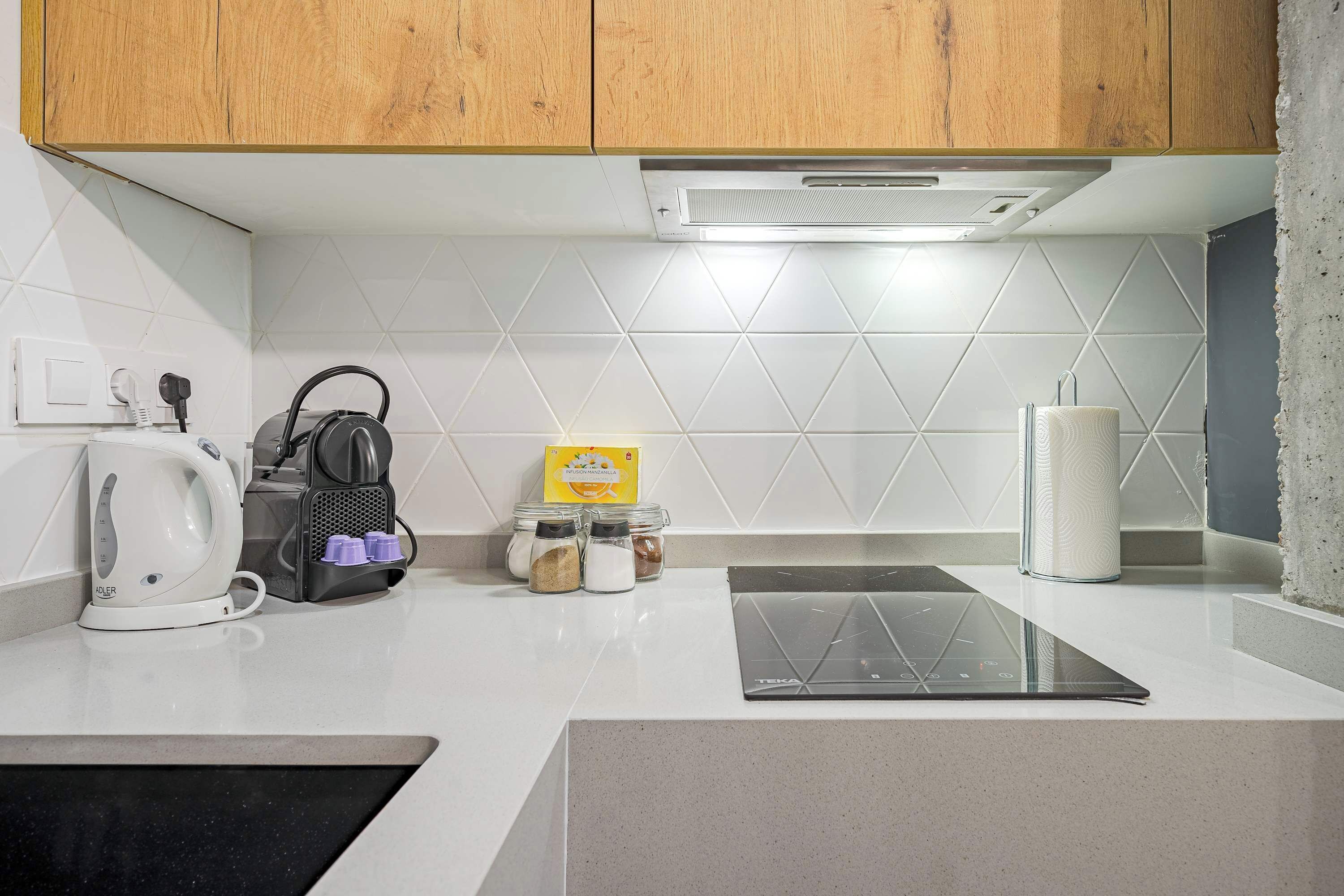 A modern kitchen with white tiles, wooden cabinets, a kettle, a coffee maker, a tablet, and jars of spices.