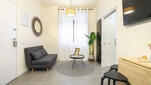 A modern living room with a grey sofa, a small round table with a picture frame, a potted plant, and a wooden dining area.