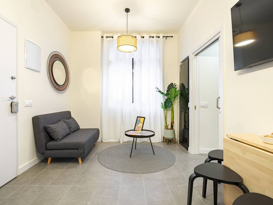 Un salon moderne avec un canapé gris, une petite table ronde sur laquelle est posé un cadre photo, une plante en pot et un coin repas en bois.