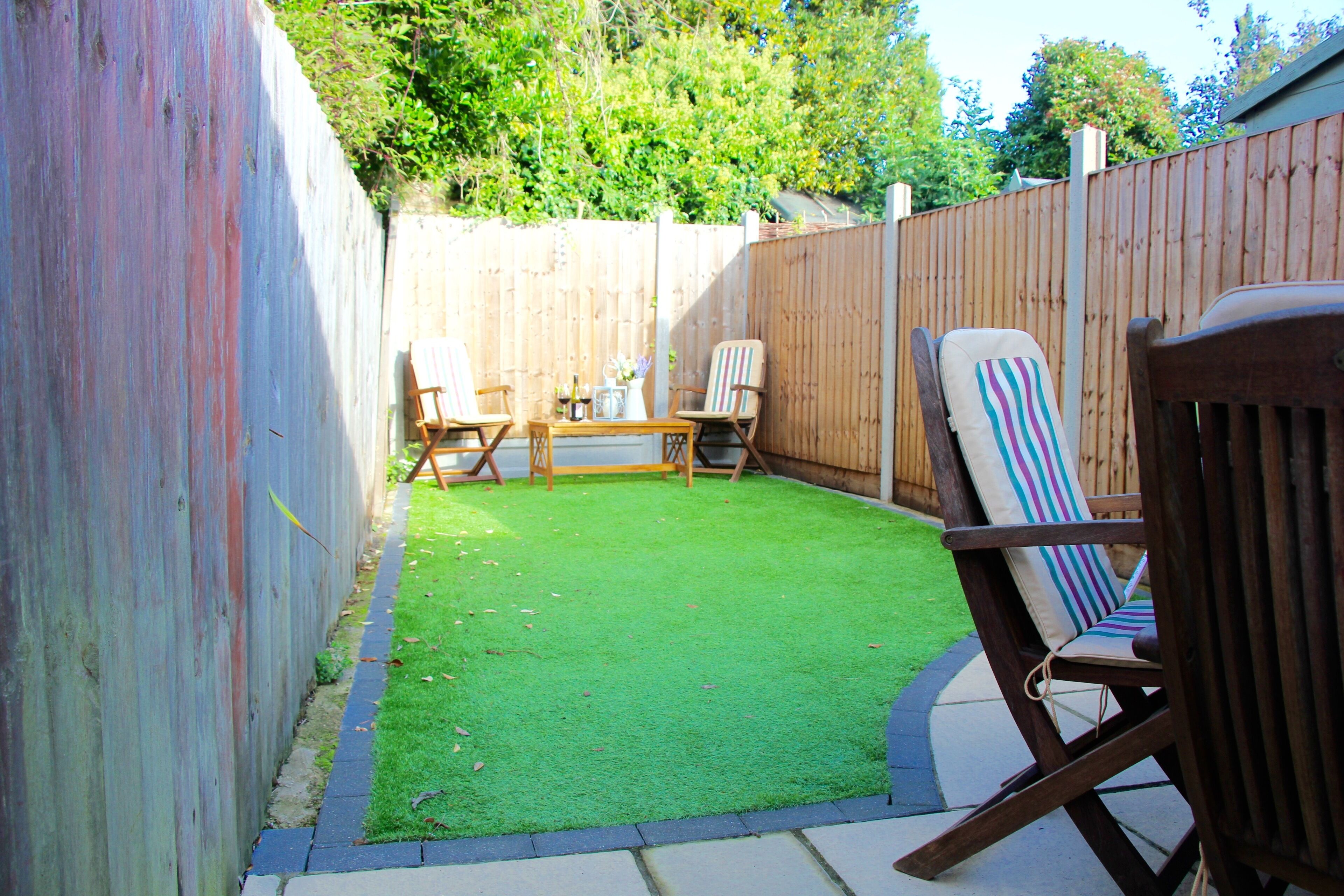 A backyard with a wooden fence, a green lawn, and two wooden chairs with cushions.