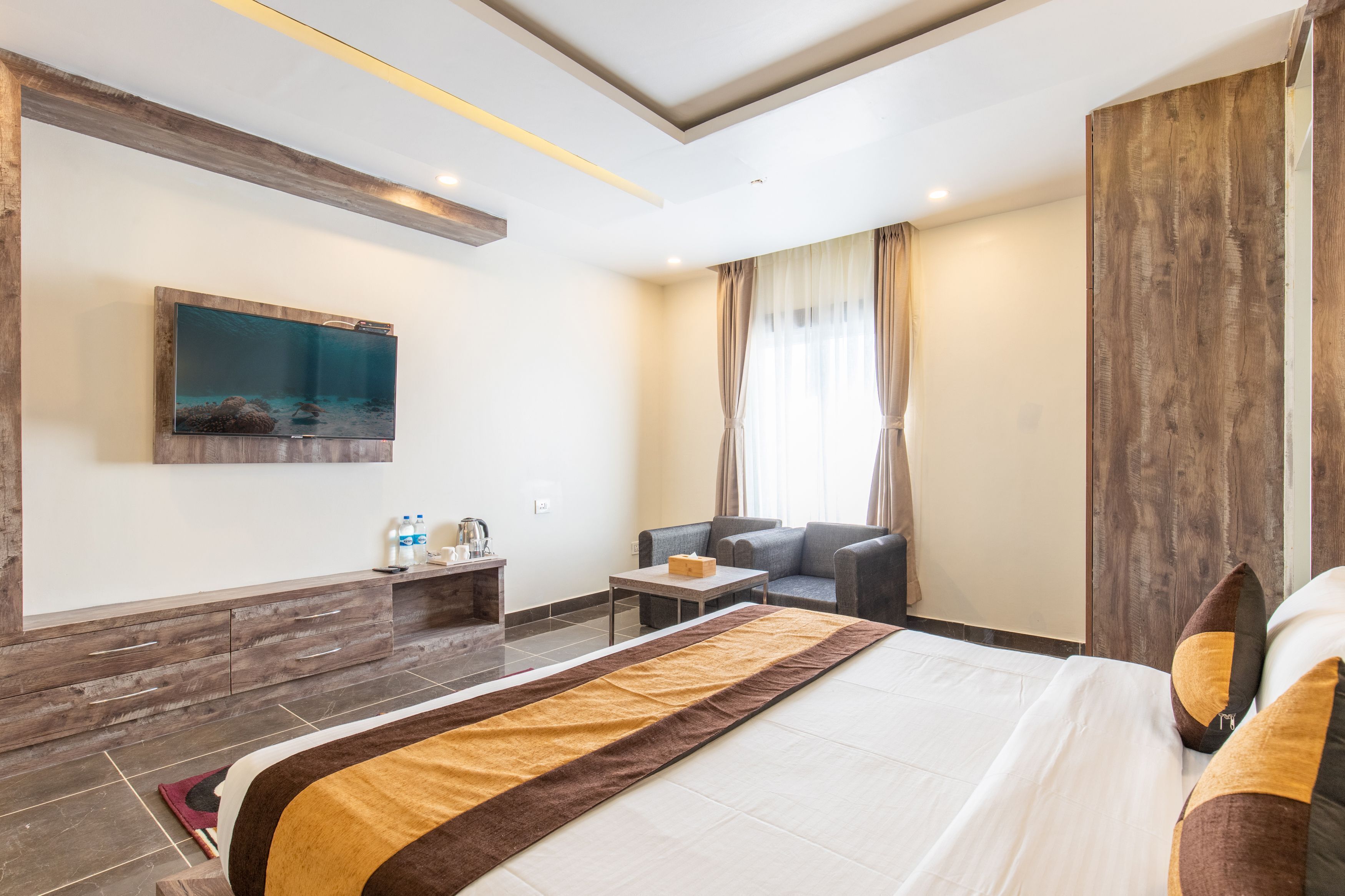 A hotel room with a large bed, a sofa, a small table, a TV mounted on the wall, and a window with curtains.