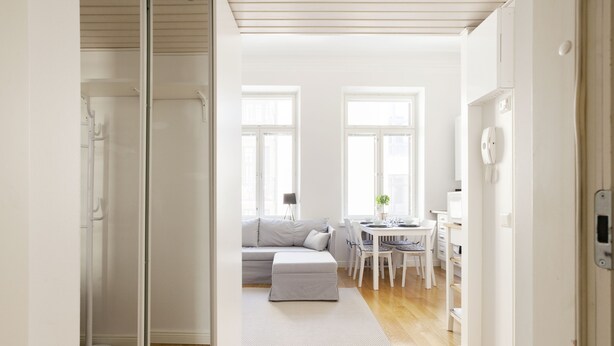 Et moderne, lyst værelse med trægulv, en grå sofa, et spisebord med stole og et hvidt køleskab.