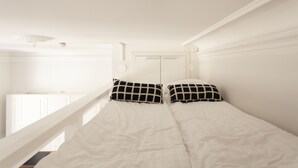 En enkeltseng med hvide sengetøj og sort-hvide ternede puder, et hvidt loft og en hvid radiator i baggrunden.