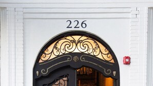 Een zwarte ijzeren deur met een decoratief ontwerp, het getal '226' erboven en een klein rood licht aan de muur rechts.
