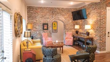 A living room with a brick wall, a flat-screen TV, a yellow sofa, patterned chairs, a wooden coffee table, and a fireplace.