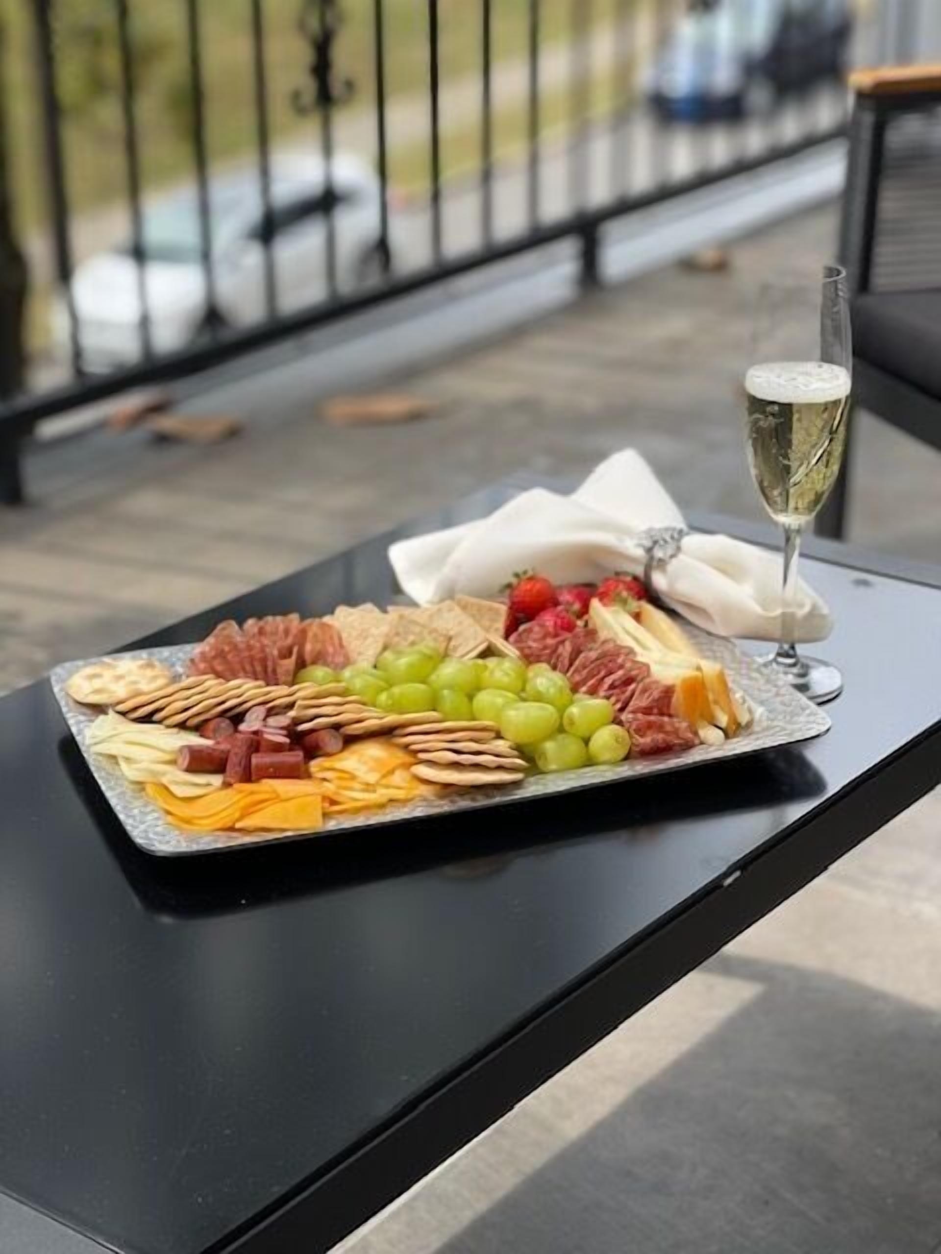 A charcuterie board with various meats, cheeses, and fruits on a tray, accompanied by a glass of champagne, placed on a table outdoors.