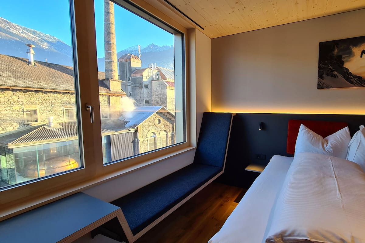 Habitación de hotel con un ventanal grande, una cama, un banco y un cuadro en la pared.