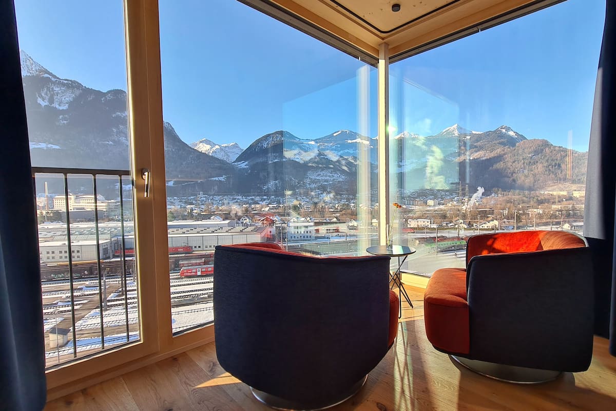 Una habitación con un ventanal que ofrece vistas a un paisaje montañoso y una estación de tren. Hay dos sillas modernas y una mesita en la habitación.