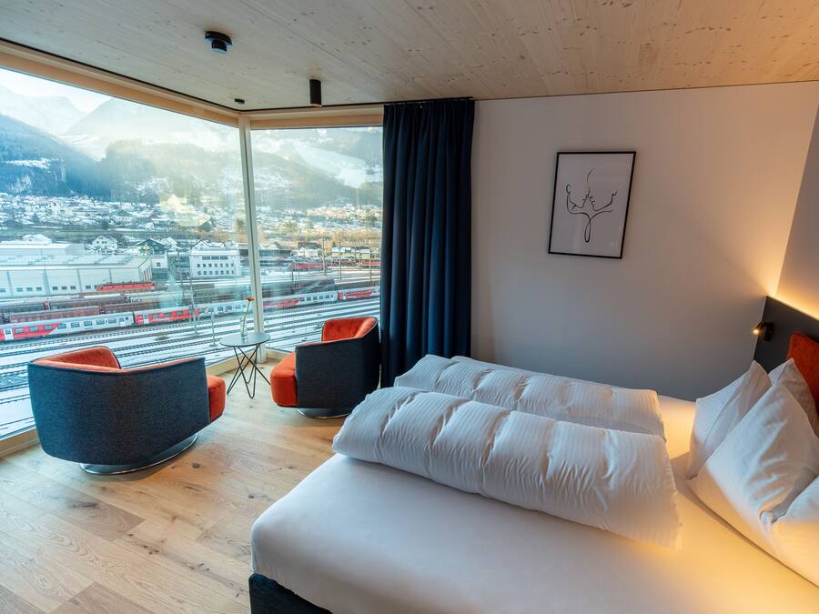 Et moderne hotelværelse med en stor seng, to lænestole, udsigt til by og bjerge, samt et billede i ramme på væggen.