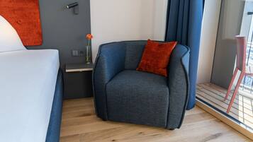 A modern hotel room with a grey sofa, a red cushion, a small table with a vase of flowers, and a window with curtains.