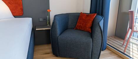 A modern hotel room with a grey sofa, a red cushion, a small table with a vase of flowers, and a window with curtains.