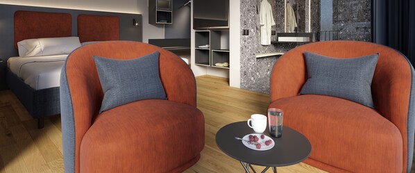 Una habitación de hotel moderna con una cama, dos sillones naranjas con cojines grises, una mesita redonda con una taza y un plato de uvas, y un baño con espejo y toallero.