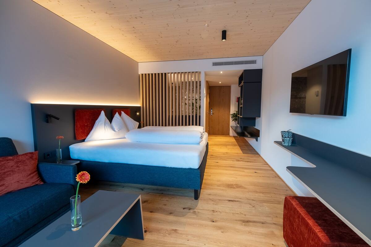 Una habitación de hotel moderna con una cama grande, un sofá, una mesa de centro con un jarrón y un televisor montado en la pared.