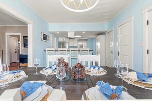 A dining area with a wooden table set for a meal, white chairs, and a kitchen in the background.