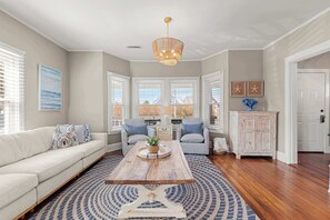 A living room with a sofa, two armchairs, a coffee table, a chandelier, and a view through large windows. - Walk to Nantucket Sound Beaches! (Dennis Port)