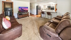 Ein Wohnzimmer mit einer braunen Ledersofa, einem Fernseher und einem Gemälde. Im Hintergrund ist eine Küche mit weißen Schränken und einem Kühlschrank zu sehen.
