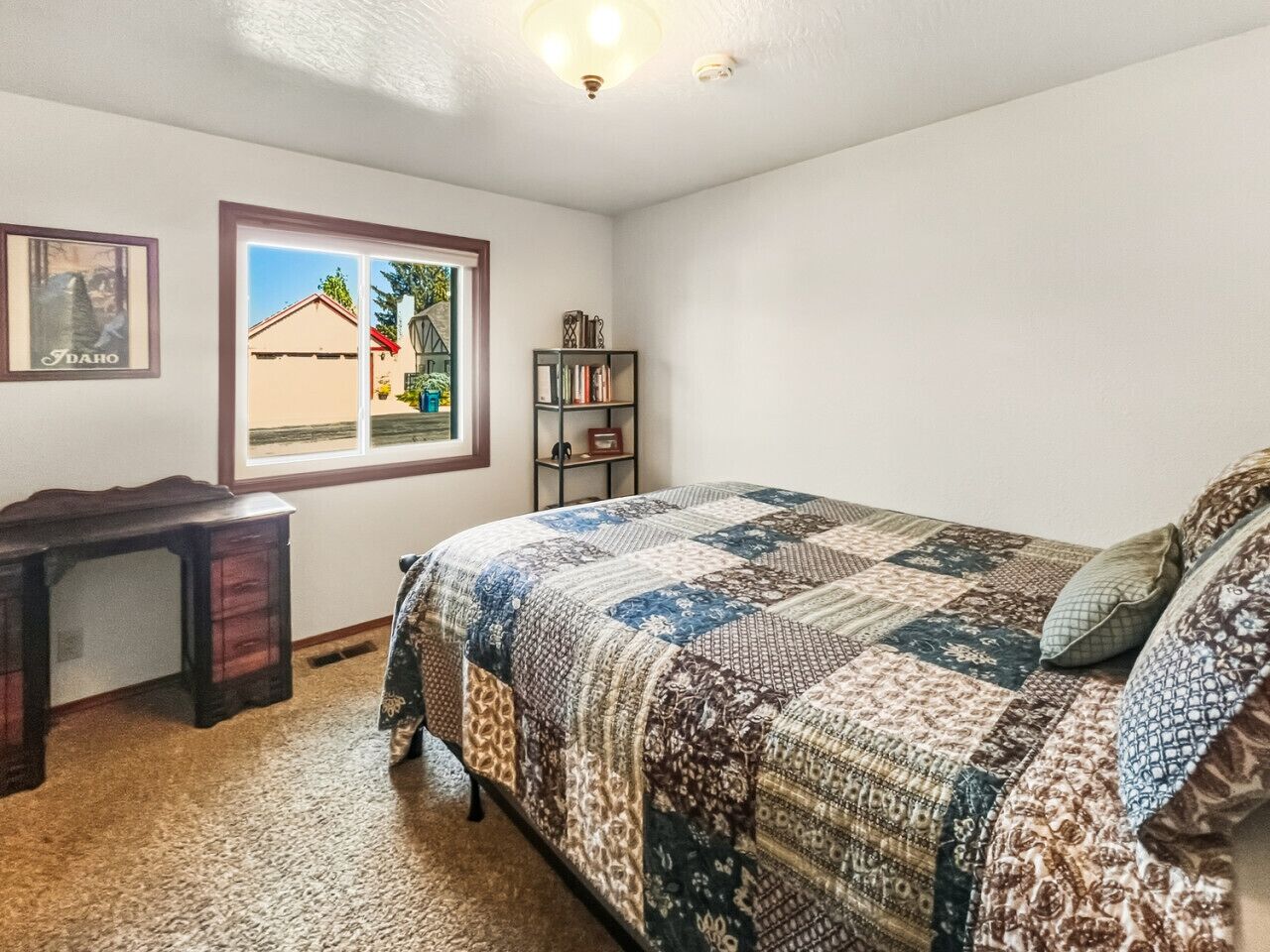 A bedroom with a bed, a desk, a bookshelf, and a window with a view of a house.