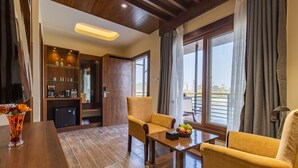 A modern living room with a wooden coffee table, two yellow armchairs, a small table with a fruit bowl, and a glass shelf with bottles. - Lumbini Palace Resort (Dayanagar)