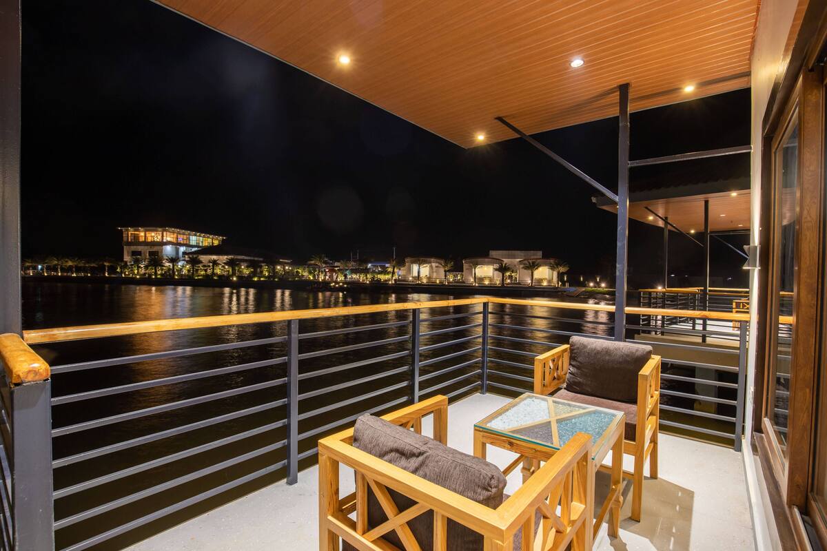 Ein Balkon mit Blick auf einen Fluss bei Nacht, ausgestattet mit Holzmöbeln und einem Geländer.