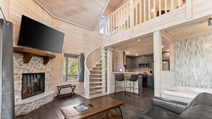 Een ruime woonkamer met een open haard, een tv bevestigd aan een houten balk, een trap naar een hoger terras, en een moderne keuken op de achtergrond.