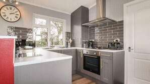 A modern kitchen with grey cabinetry, a stainless steel range hood, and a built-in oven. - Sycamore Lodge (Helston)