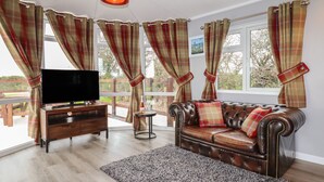 A living room with a leather sofa, a TV on a wooden stand, a small table with a bottle and glasses, plaid curtains, and a view of the outdoors. - Sycamore Lodge (Helston)