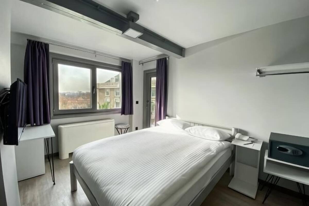 A modern hotel room with a large bed, a desk, a chair, and a view of the city through a window.