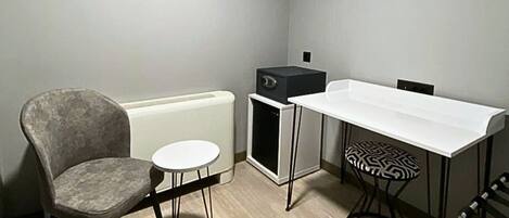 A small, compact room with a grey upholstered chair, a small round table, a white table, a black and white patterned stool, and a black appliance unit.