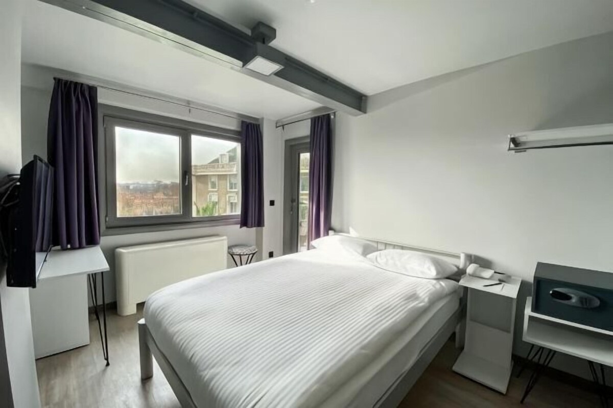 Et moderne hotelværelse med en stor seng, et skrivebord, en stol og udsigt over byen gennem et vindue.