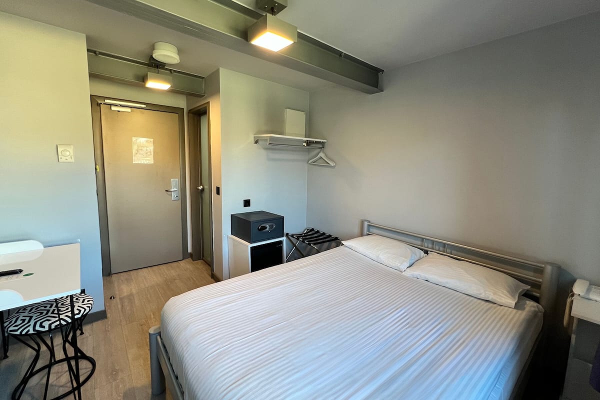 A hotel room with a bed, bedside table, and a desk with a chair. There is a coat rack and a small cabinet.
