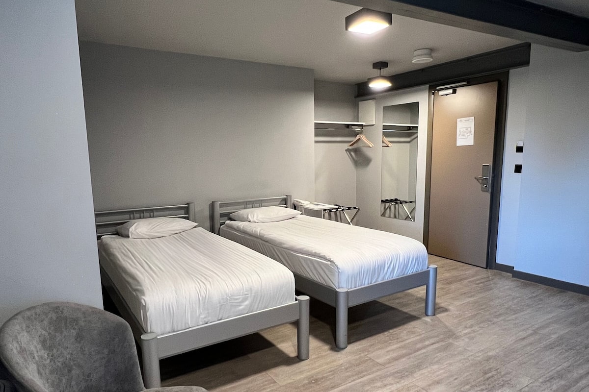 A hotel room with two single beds, a chair, a mirror, and a door.